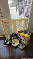 Photo of pet gate, training pads, door hook, fabric bag and two dog bowls on the floor near a door.