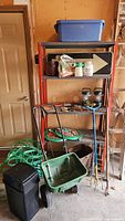 Overview of shelving unit in garage with garden tools, seed spreader, hoses, and containers arranged on and around it.