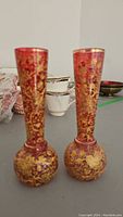 Pair of cranberry glass vases with detailed gold handpainted leaf and vine designs paired side-by-side showing full height and design.