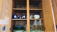 Cabinet with shelves holding 6 clear wine glasses, multiple green and blue plates, and 5 blue mugs along with a wine bottle holder.