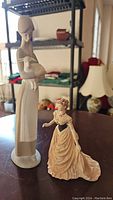 Photo of Coalport Beau Monde Molly 5.5-inch figurine next to NAO porcelain bisque lady with lamb 10.5-inch figurine on a wood table with shelving and home decor background.