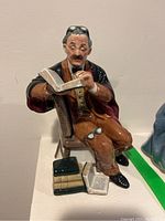 Front angle of the Royal Doulton 'The Professor' figurine showing the seated older male figure holding an open book, glasses on his head and knee, and books scattered at his feet.
