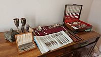 Wide view of all items from the set laid out on a wooden sideboard including silver goblets, silver nutcracker, salt cellars, multiple silver spoons and forks, and steak knives in a wood case with red interior.