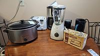 Group photo of all small appliances on a wooden surface including crockpot, toaster, blender, coffee maker, portable mixer in box, kettle and coffee grinder