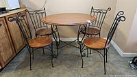 Full view of round laminate top table with wrought iron base and four wrought iron chairs with wooden seats around it in a room corner.