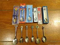 Full view photo showing all 12 collector spoons arranged in a row, five in original packaging, seven loose with varied design and metal finishes, mostly silver plated.