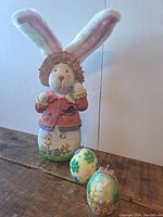 Front view of the large decorative Easter bunny figurine with fabric ears, holding a small egg, and two egg-shaped candles in front of it.
