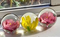 Three glass paperweights with embedded flower designs placed on windowsill: two with pink flowers and one with yellow flower inside clear glass spheres.