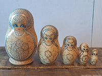 Front view of all five traditional Russian stacking dolls arranged in descending size order on a wooden surface, showing detailed facial features and floral motifs in beige and silver.