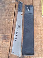 Machete alongside its black nylon sheath displayed on wooden surface.