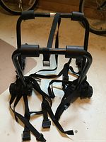 Bike carrier frame showing black metal bars and plastic components with securing straps on floor.