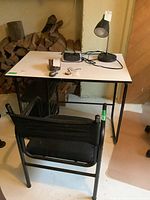 Desk with white composite top, black metal frame and two mesh storage shelves below; black fabric and metal chair in front.