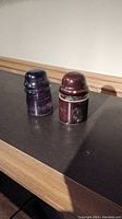 Photo of two electrical insulators on a dark surface: one glass, purple-tinted with ribbed texture, the other reddish-brown ceramic with grooved ring near top.
