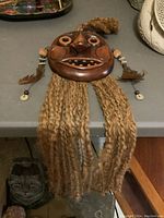 Front view of the wooden tribal mask showing facial carving, natural fiber hair hanging, and beaded, feathered braided hair attachments on sides.