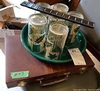 Top view of the tray with six tumblers with leaf motif and the brown portable backgammon case underneath, a ruler laid across for scale.