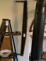 Vintage wood handled buck saw hanging on wall with large toothed blade and wooden handles visible, next to a wooden ladder and other items.