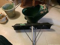Photo showing green tin watering can with floral sunflower design and black metal roof shovel without handle placed on the floor.