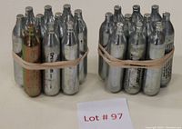 Two bundles of 10 silver CO2 cartridges each, held together by rubber bands, arranged standing upright on a surface. One cartridge shows discoloration.