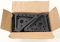 Two black triangular rubber wheel chocks inside a cardboard box, showing the side with holes and ridged gripping surface.