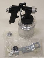 Front view of sprayer gun with attached metal paint container and additional parts in bags.