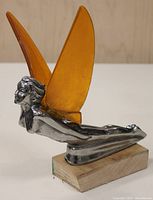 Side view of metal angel hood ornament with yellow translucent wings laying horizontally on wooden base.