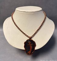 Copper-tone necklace displayed on bust showing chain and pendant with stylized figure design.