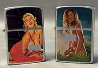 Two chrome-tone metal tobacco lighters with colorful risqué pin-up images side by side on neutral surface