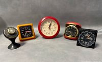 Five vintage travelling alarm clocks in a row with front faces visible, showing different shapes and colors including red, yellow, black, and leather casing.