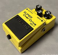 Side angled view showing the pedal's yellow housing, input and output jack labels, and three black control knobs with silver tops.