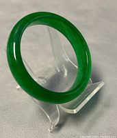 Polished green jade bangle displayed on clear stand, showing smooth rounded shape and rich translucent green color.