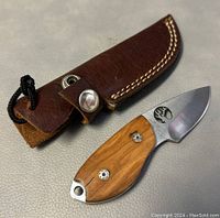 Mini hunter's knife with wooden handle and stainless stainless steel blade, next to brown leather sheath with snap closure, white stitching visible