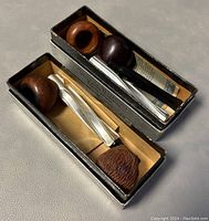 Two vintage tobacco pipes inside their metal boxes, showing different carved wooden bowls and black mouthpieces.
