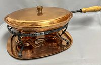 Copper pan on warmer stand with copper tray underneath, showing wooden handle and brass knob on lid