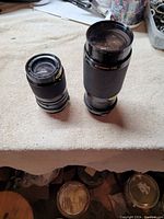 Two black vintage camera lenses on a beige cloth surface, one smaller 85-135mm lens and one larger 80-200mm lens.