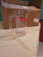Single clear plastic Budweiser pitcher displayed upright next to a cardboard box.