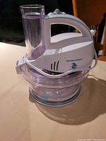 Front view of white Hamilton Beach electric grater with transparent bowl and food feed tube, sitting on a table against a wooden chair background.