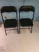 Two black metal folding chairs with padded vinyl seats and backs placed on carpet against a light blue wall.