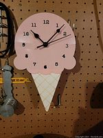 Wall clock shaped like an ice cream cone with pink scoop and beige cone, mounted on pegboard background.