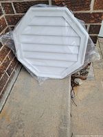 White octagonal decorative gable vent in original plastic packaging, leaning against brick wall outdoors.