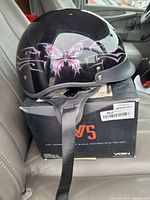 New glossy black Vcan V5 motorcycle helmet with pink butterfly design placed on its original box on car seat