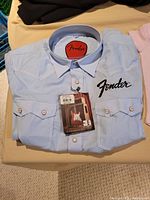 Folded men's small light blue button-up shirt with Fender logo on front pocket and tag attached.