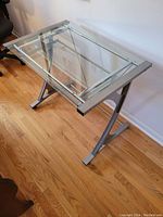 Clear view of the desk showing glass top, metal X-shaped legs, and sliding keyboard shelf, standing on hardwood floor.