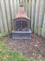 Outdoor metal fireplace with chimney, black and rust-brown finish, placed outside on soil against a wooden fence.