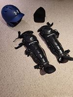 Blue Under Armour batting helmet, black catcher shin guards, and black cap displayed together on carpet