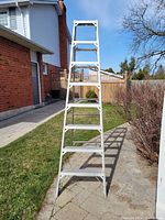 Front view of an 8-foot aluminum step ladder standing upright on a paved surface near a brick wall and grassy area.