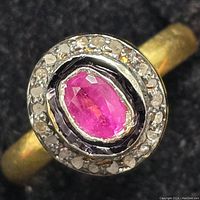 Close-up photo showing the silver ring with a central oval ruby and diamond halo under direct light on a dark fabric background.