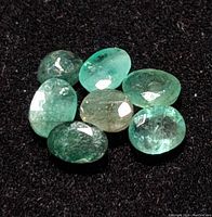 Seven loose oval-shaped emerald gemstones displayed on black fabric, showing natural inclusions and color variations from pale to medium green.