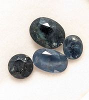 Four genuine sapphire gemstones displayed on a light background. Includes two oval and two round faceted stones of varying sizes and blue shades.