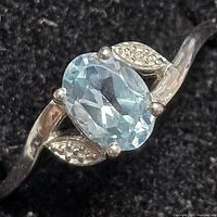 Close-up front view of silver ring featuring an oval light blue faceted topaz gemstone with decorative ribbed silver leaf accents on either side, set on a polished silver band.