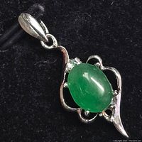 Close-up front view of silver pendant with carved design holding green jadeite oval stone and several small clear accent stones.